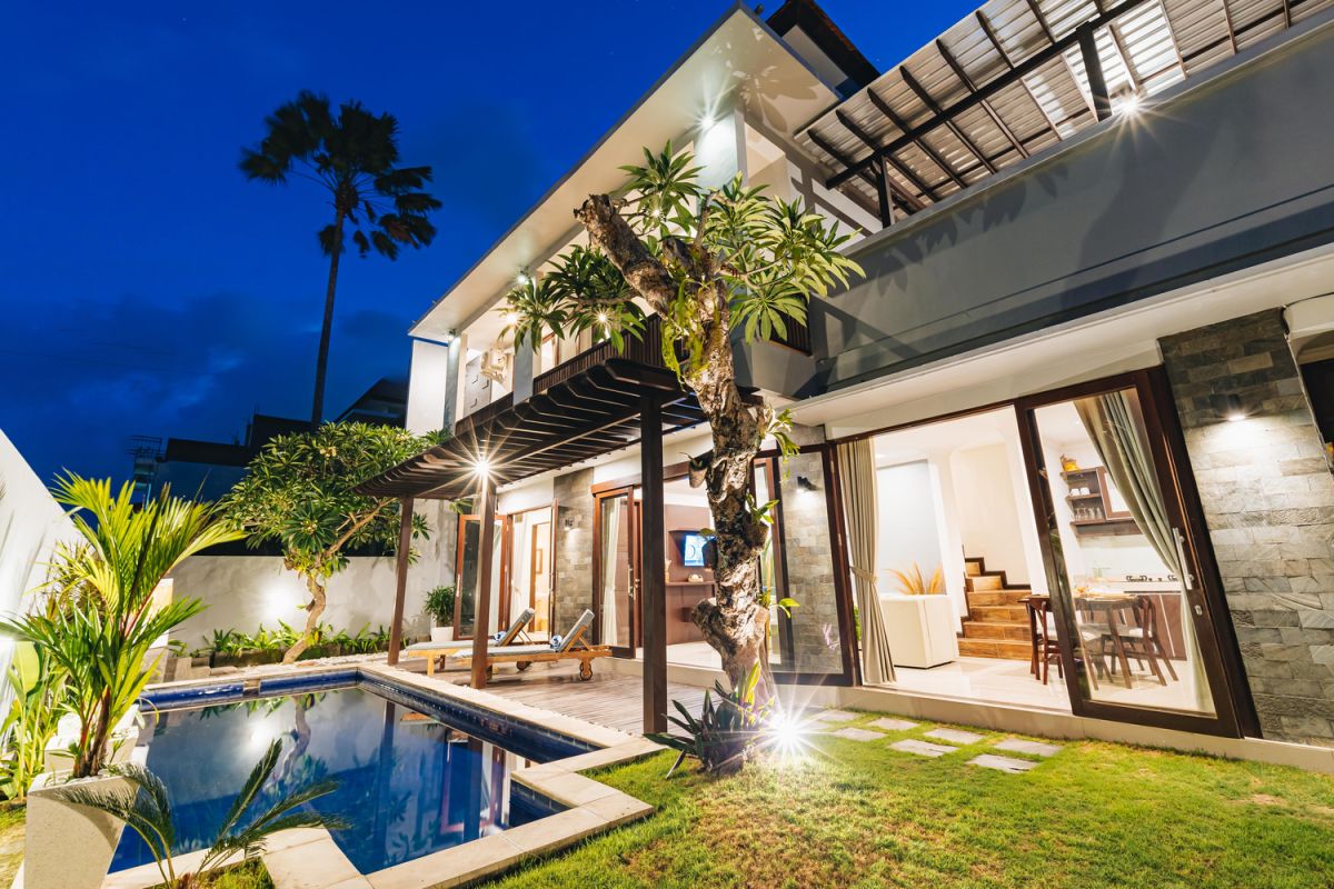 Modern Balinese living room and dining area with beige sofas, wooden table set for meal, kitchen counter, potted plants, and stairs. - Bali Villas