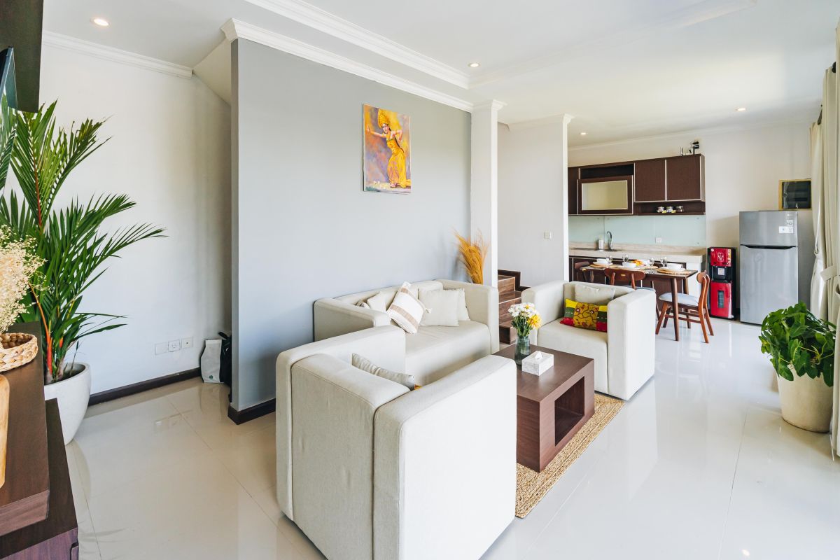 Modern Balinese living room and dining area with beige sofas, wooden table set for meal, kitchen counter, potted plants, and stairs. - Bali Villas