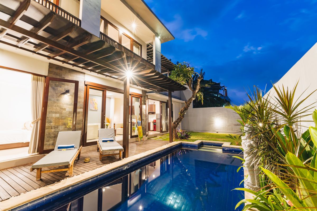 Modern Balinese living room and dining area with beige sofas, wooden table set for meal, kitchen counter, potted plants, and stairs. - Bali Villas