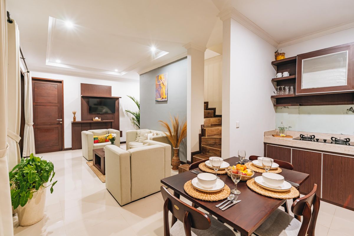 Modern Balinese living room and dining area with beige sofas, wooden table set for meal, kitchen counter, potted plants, and stairs. - Bali Villas