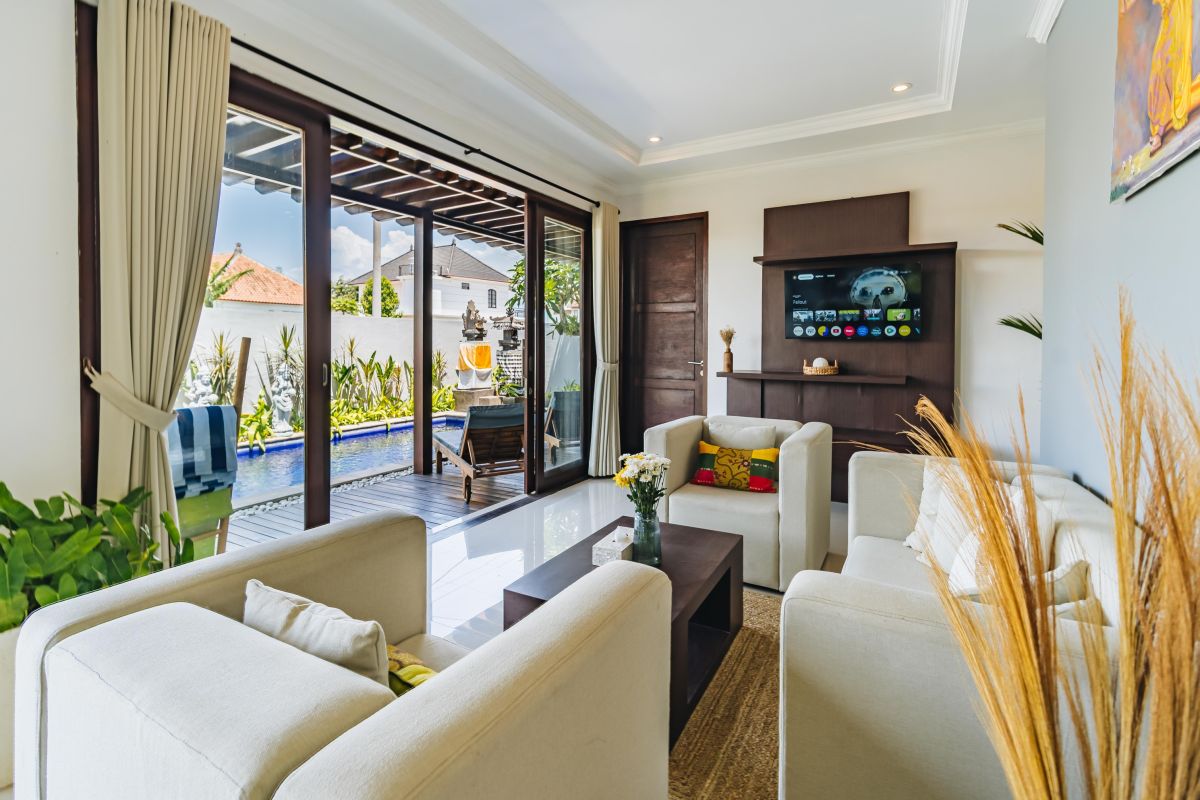 Modern Balinese living room and dining area with beige sofas, wooden table set for meal, kitchen counter, potted plants, and stairs. - Bali Villas