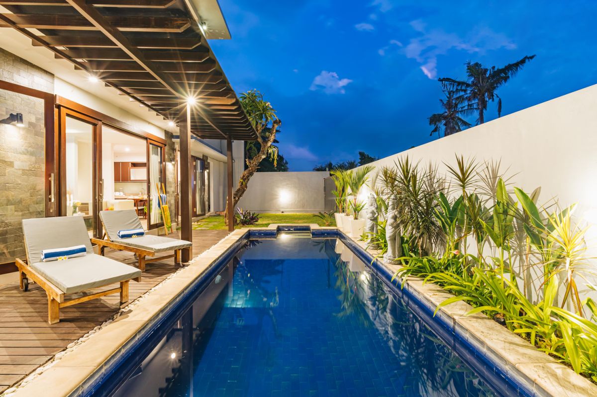 Modern Balinese living room and dining area with beige sofas, wooden table set for meal, kitchen counter, potted plants, and stairs. - Bali Villas