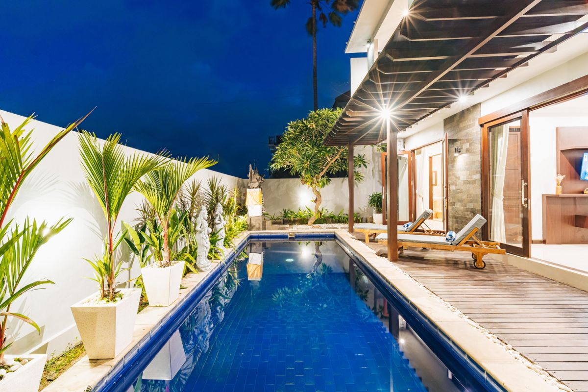 Modern Balinese living room and dining area with beige sofas, wooden table set for meal, kitchen counter, potted plants, and stairs. - Bali Villas
