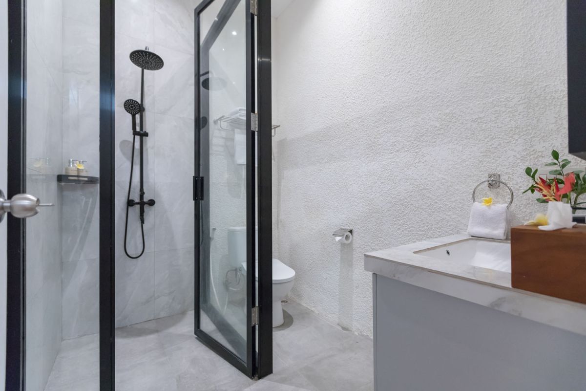 Modern bathroom with black-framed glass shower enclosure, rainfall showerhead, white toilet, and marble vanity sink. - Bali Villas