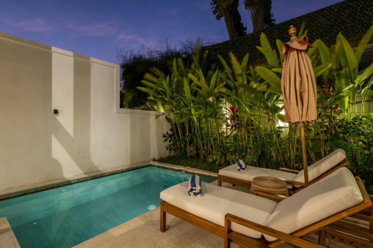 Serene tropical infinity pool at dusk with white lounge chairs, beige umbrella, and lush banana palms under soft lighting. - Bali Villas