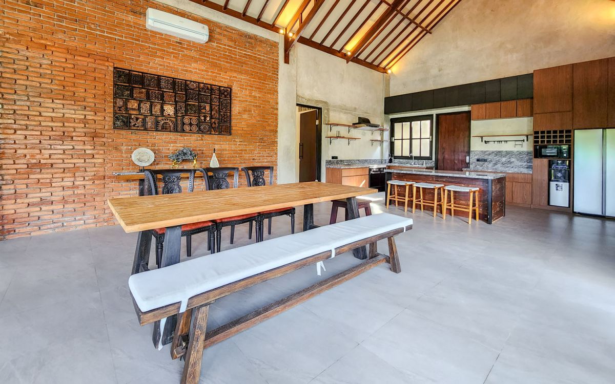 Spacious Balinese-style living room with gray sofa, wooden coffee table, patterned armchair, abstract sculpture, tropical plants, and infinity pool view through floor-to-ceiling windows. - Bali Villas