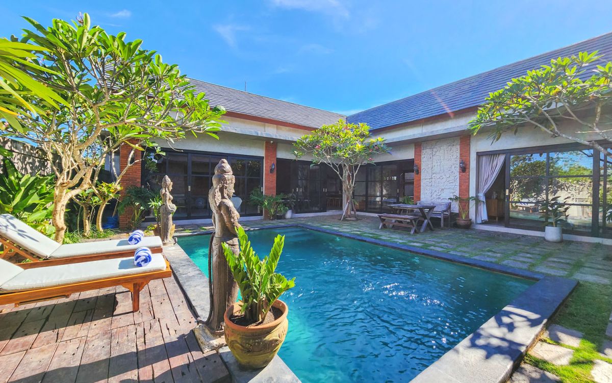 Spacious Balinese-style living room with gray sofa, wooden coffee table, patterned armchair, abstract sculpture, tropical plants, and infinity pool view through floor-to-ceiling windows. - Bali Villas