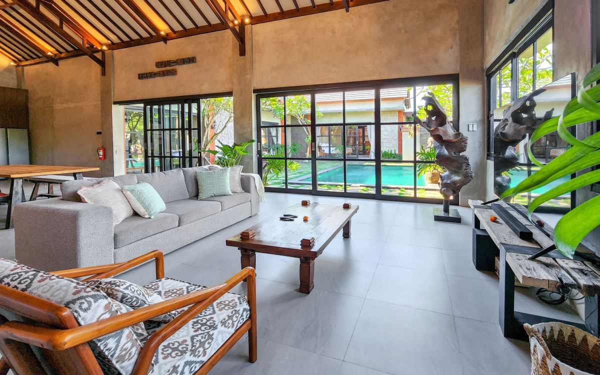 Spacious Balinese-style living room with gray sofa, wooden coffee table, patterned armchair, abstract sculpture, tropical plants, and infinity pool view through floor-to-ceiling windows. - Bali Villas