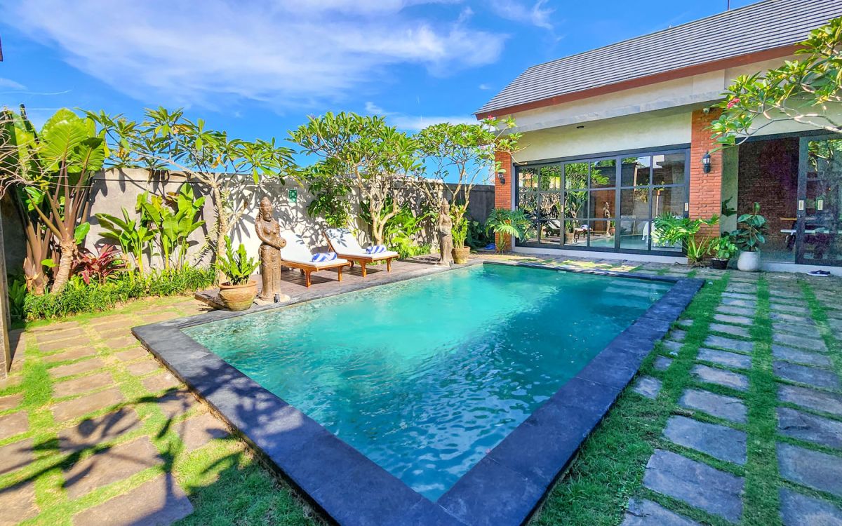 Spacious Balinese-style living room with gray sofa, wooden coffee table, patterned armchair, abstract sculpture, tropical plants, and infinity pool view through floor-to-ceiling windows. - Bali Villas