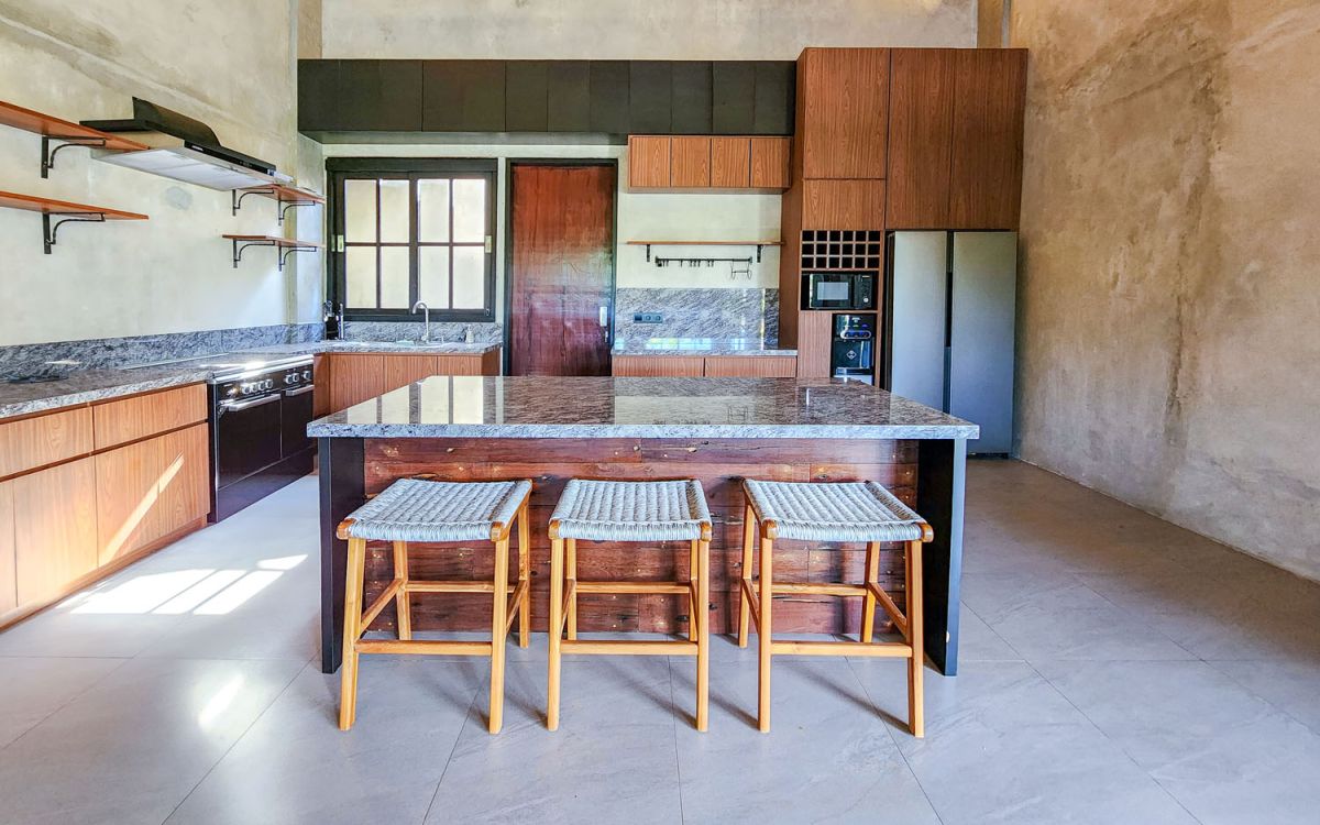 Spacious Balinese-style living room with gray sofa, wooden coffee table, patterned armchair, abstract sculpture, tropical plants, and infinity pool view through floor-to-ceiling windows. - Bali Villas