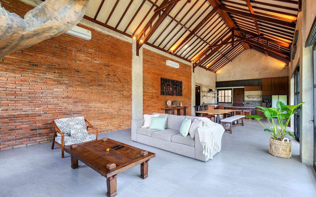 Spacious Balinese-style living room with gray sofa, wooden coffee table, patterned armchair, abstract sculpture, tropical plants, and infinity pool view through floor-to-ceiling windows. - Bali Villas