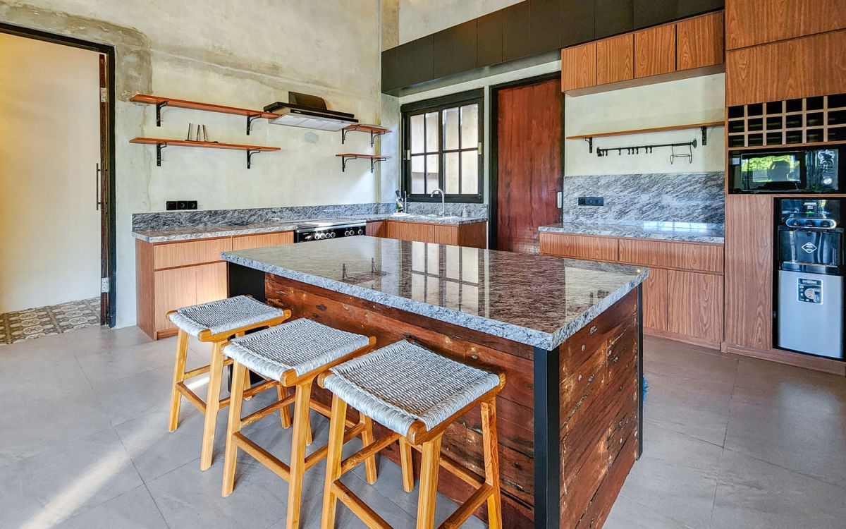 Spacious Balinese-style living room with gray sofa, wooden coffee table, patterned armchair, abstract sculpture, tropical plants, and infinity pool view through floor-to-ceiling windows. - Bali Villas