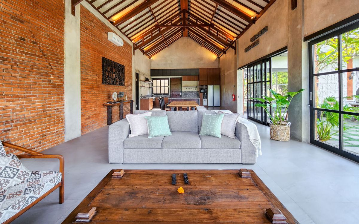 Spacious Balinese-style living room with gray sofa, wooden coffee table, patterned armchair, abstract sculpture, tropical plants, and infinity pool view through floor-to-ceiling windows. - Bali Villas