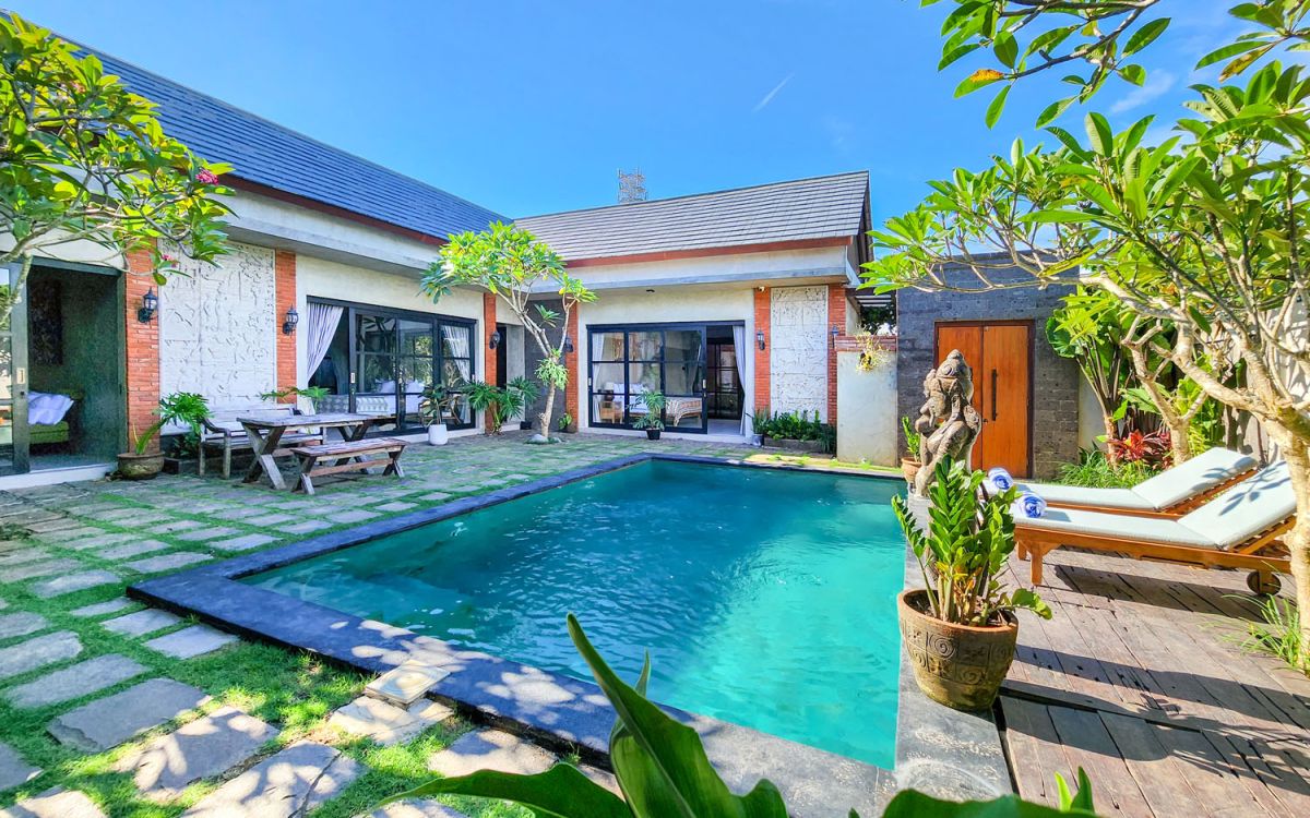 Modern Balinese-style villa with private turquoise swimming pool, wooden lounge chairs, potted plants, and lush frangipani trees in a sunny courtyard. - Bali Villas