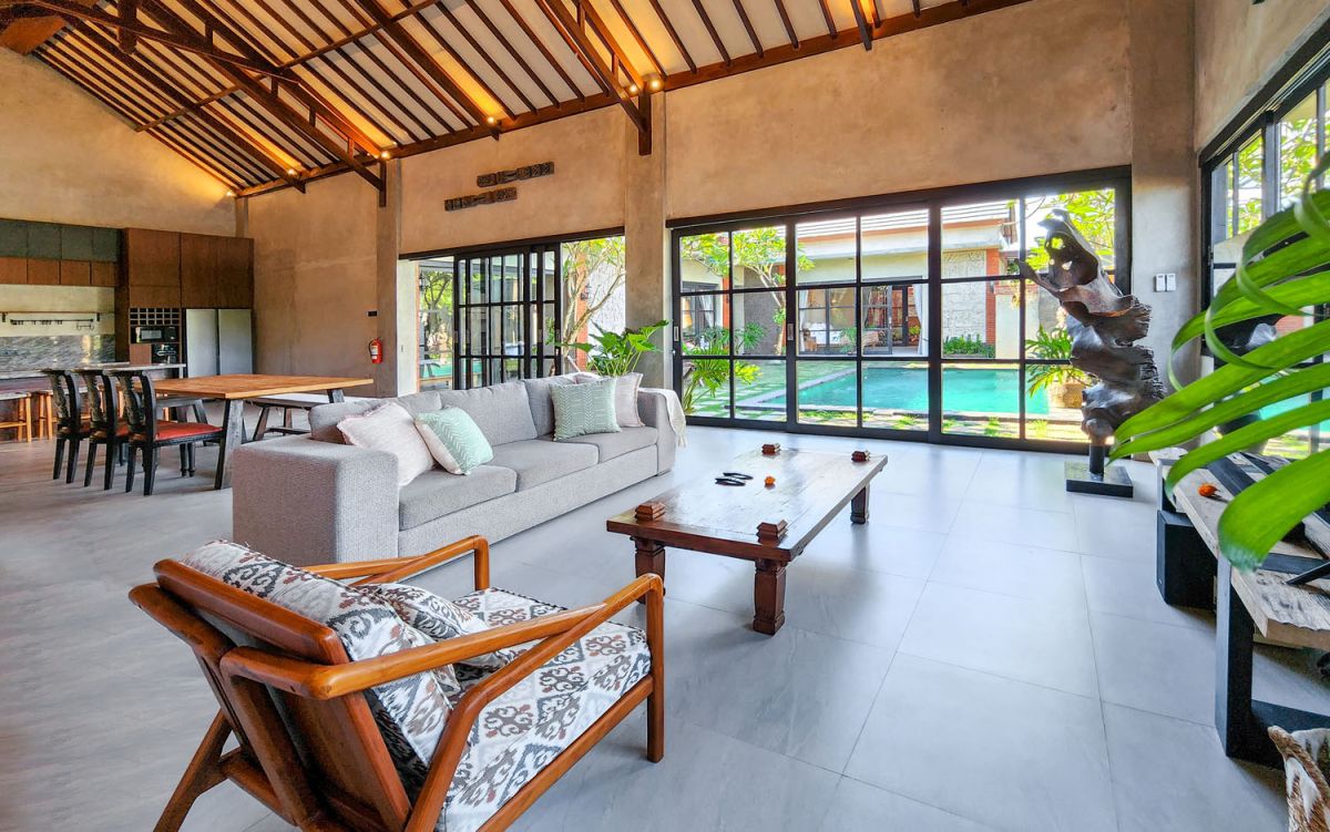 Spacious Balinese-style living room with gray sofa, wooden coffee table, patterned armchair, abstract sculpture, tropical plants, and infinity pool view through floor-to-ceiling windows. - Bali Villas