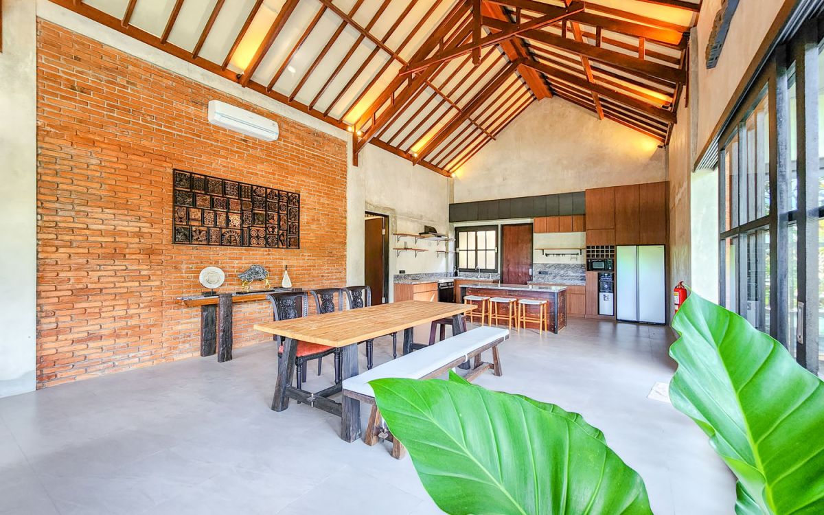 Spacious Balinese-style living room with gray sofa, wooden coffee table, patterned armchair, abstract sculpture, tropical plants, and infinity pool view through floor-to-ceiling windows. - Bali Villas