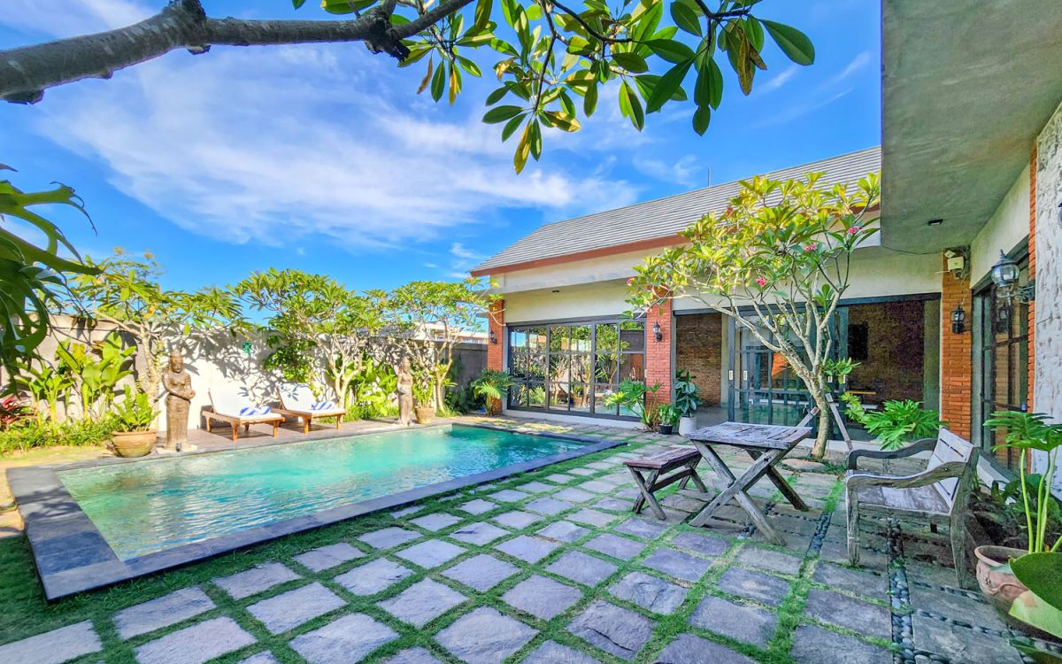 Spacious Balinese-style living room with gray sofa, wooden coffee table, patterned armchair, abstract sculpture, tropical plants, and infinity pool view through floor-to-ceiling windows. - Bali Villas