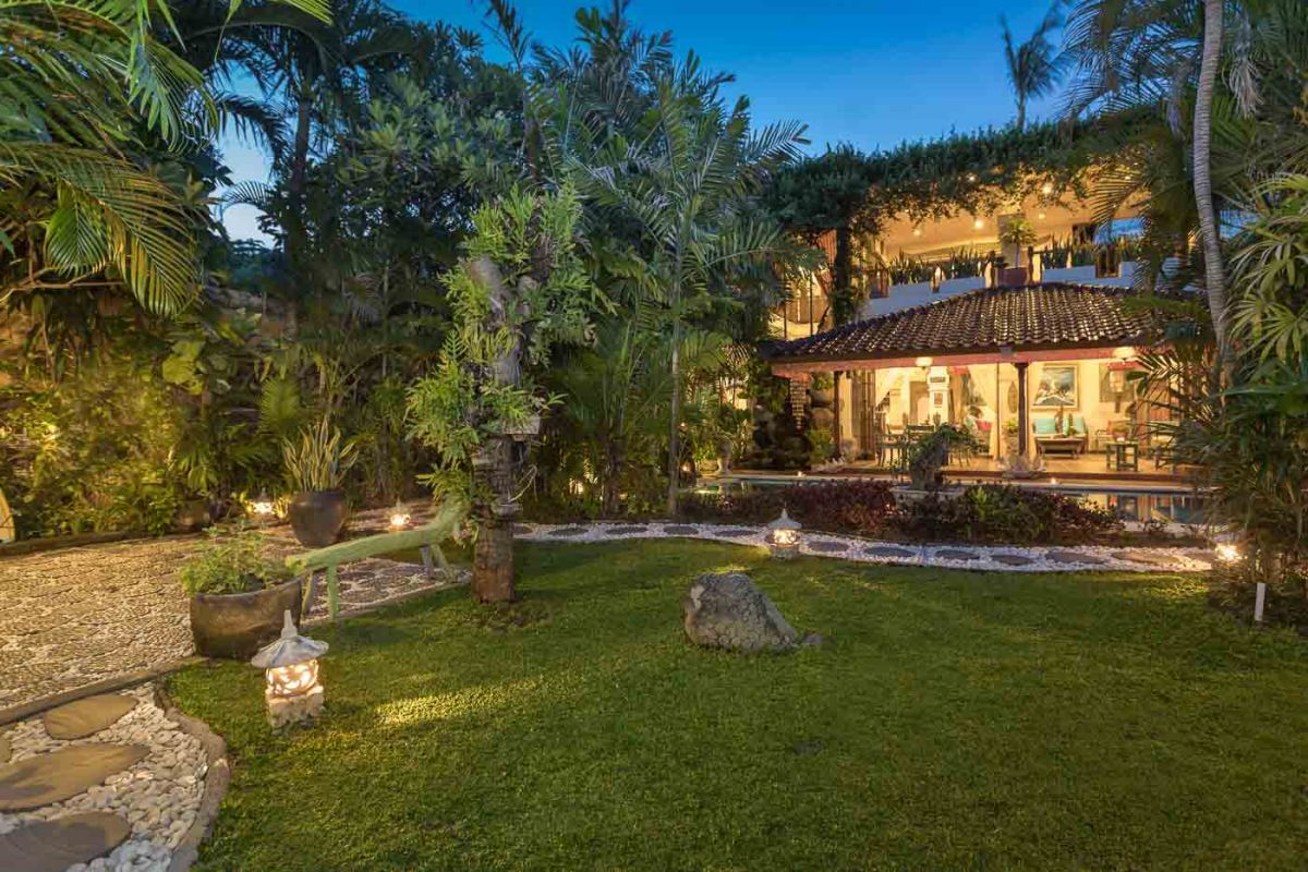 Luxurious Bali-style spa bathroom with rose-petal-filled bathtub, double black stone sinks, wooden vanities, tropical plants, and natural light from skylight. - Bali Villas