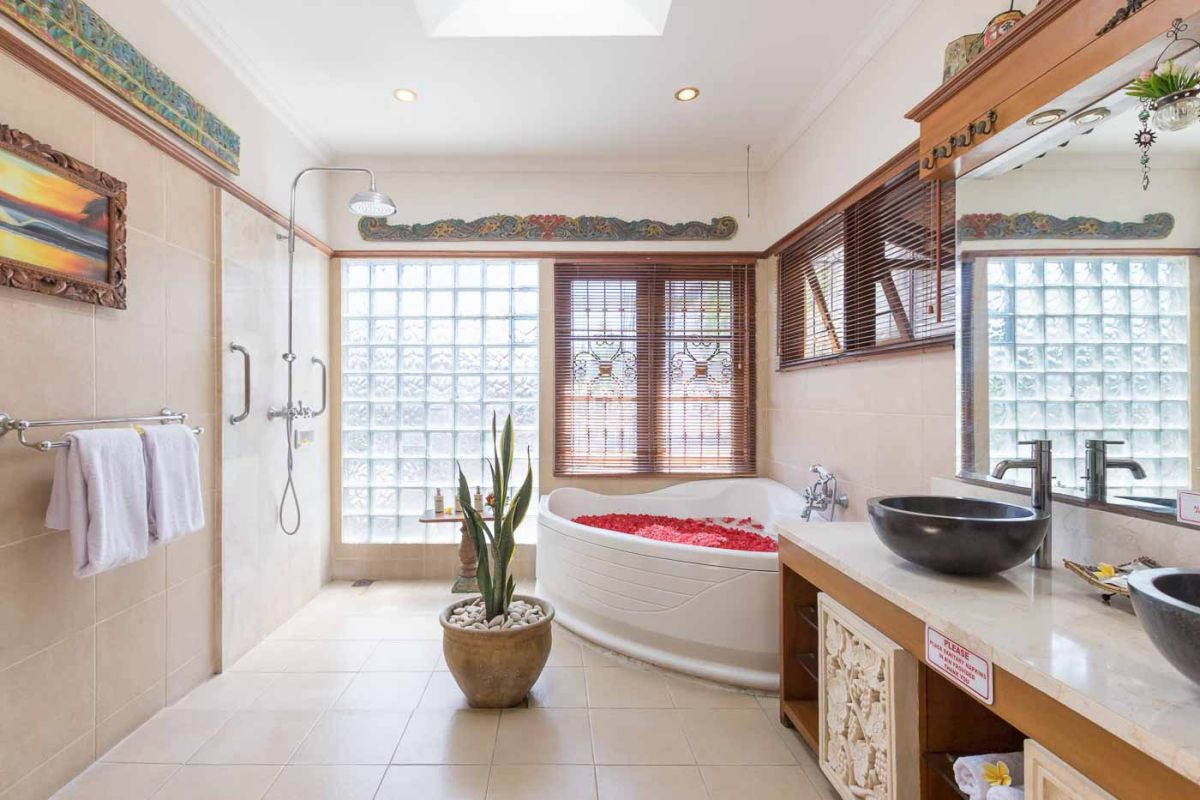Luxurious Bali-style spa bathroom with rose-petal-filled bathtub, double black stone sinks, wooden vanities, tropical plants, and natural light from skylight. - Bali Villas