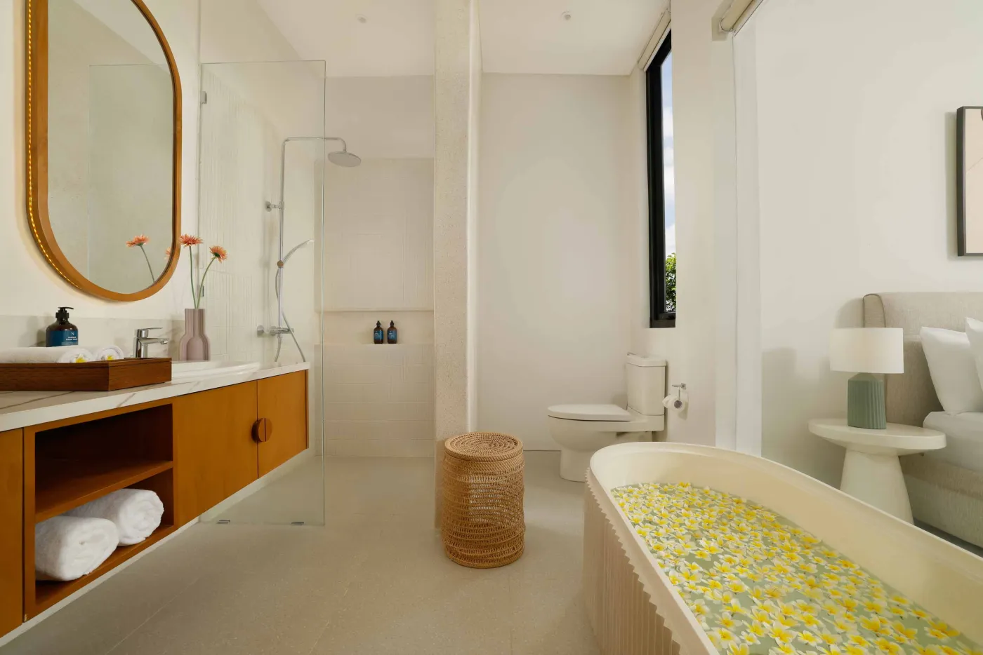 Modern minimalist bathroom with yellow flower-filled bathtub, wooden vanity, glass shower enclosure, and potted greenery. - Bali Villas