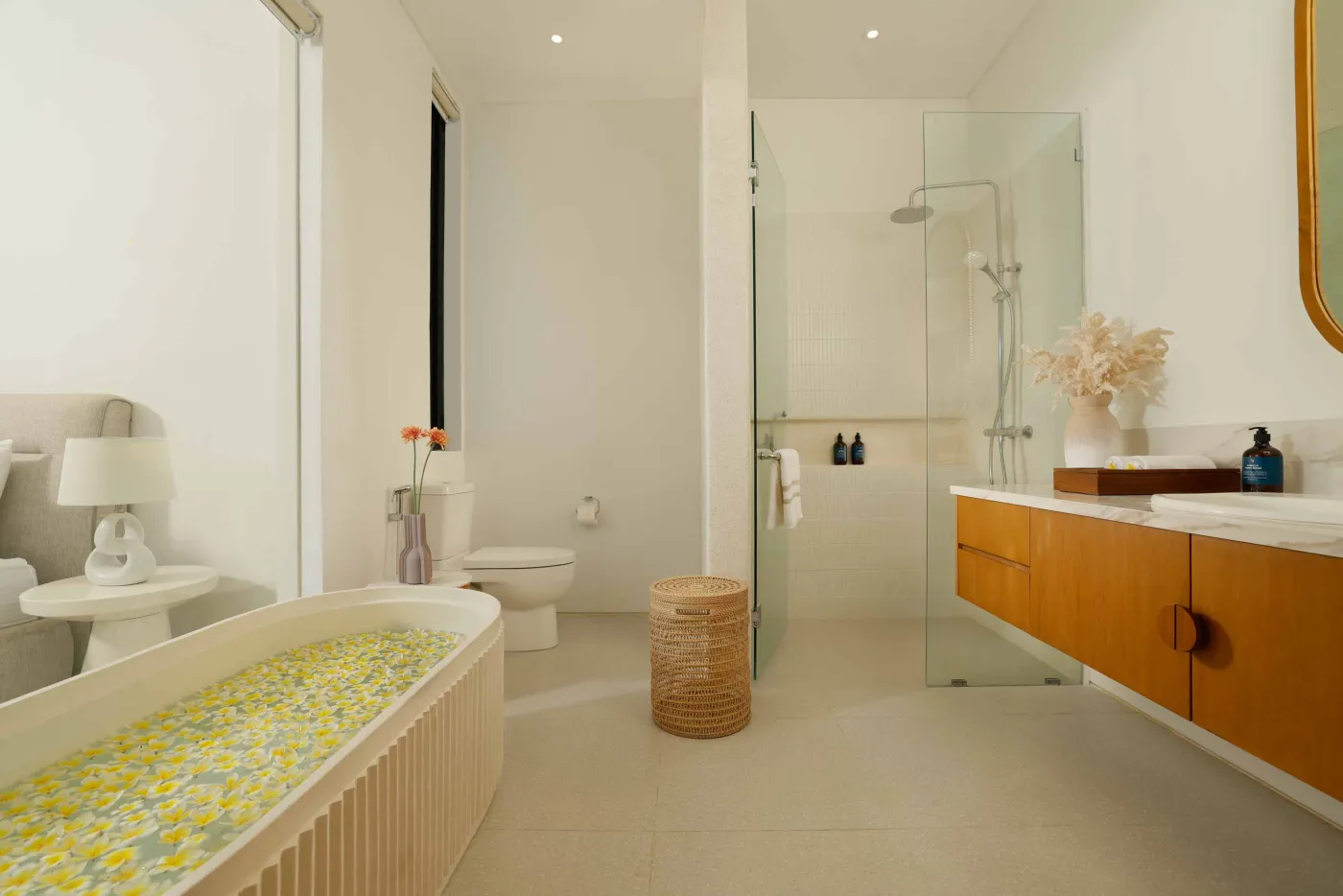 Modern minimalist bathroom with yellow flower-filled bathtub, wooden vanity, glass shower enclosure, and potted greenery. - Bali Villas