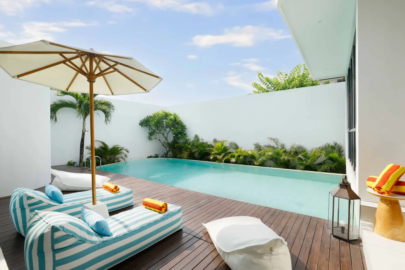 Luxurious tropical poolside retreat with turquoise infinity pool, striped lounge chairs, orange towels, and palm trees on a wooden deck. - Bali Villas