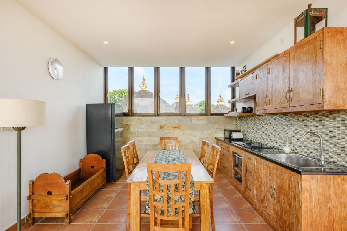 Rustic Balinese villa kitchen and dining room with wooden cabinets, stone walls, and views of traditional temple roofs. - Bali Villas