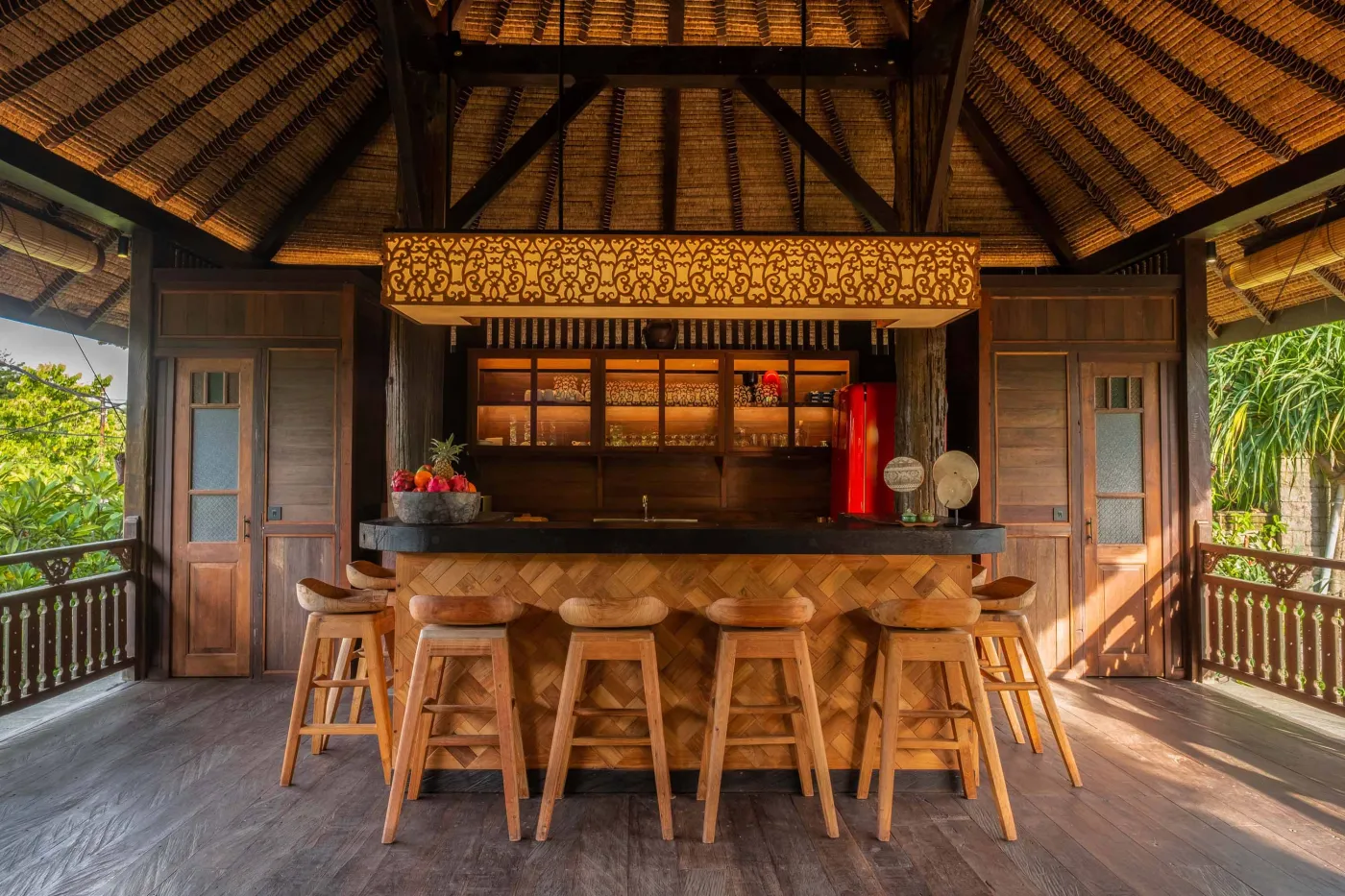 Open-air tropical resort bar with carved wooden counter, thatched roof, backlit liquor shelves, fresh fruits, and surrounding lush greenery. - Bali Villas