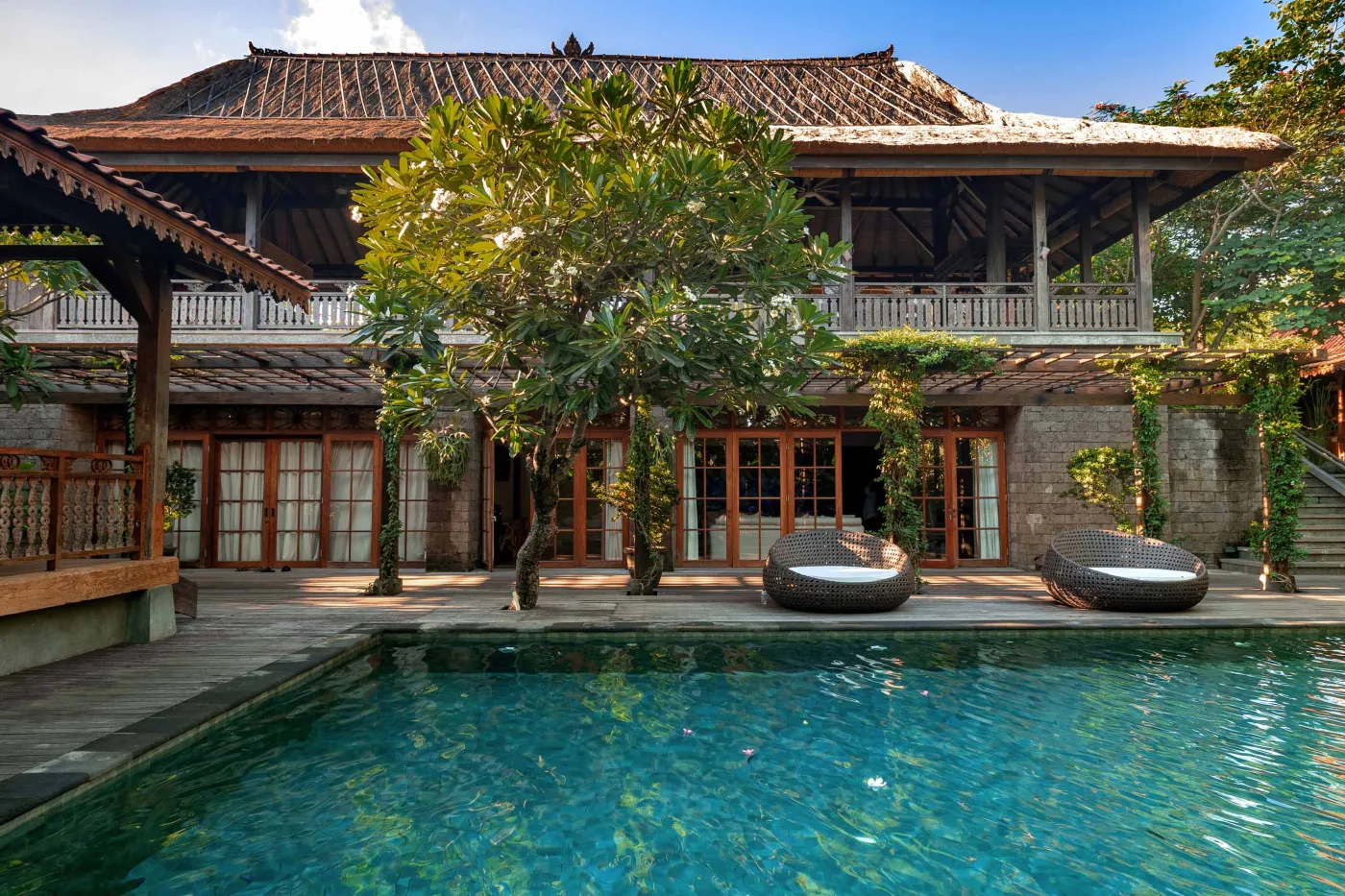 Luxurious Balinese villa with turquoise infinity pool, wooden deck, lounge chairs, and tropical garden. - Bali Villas