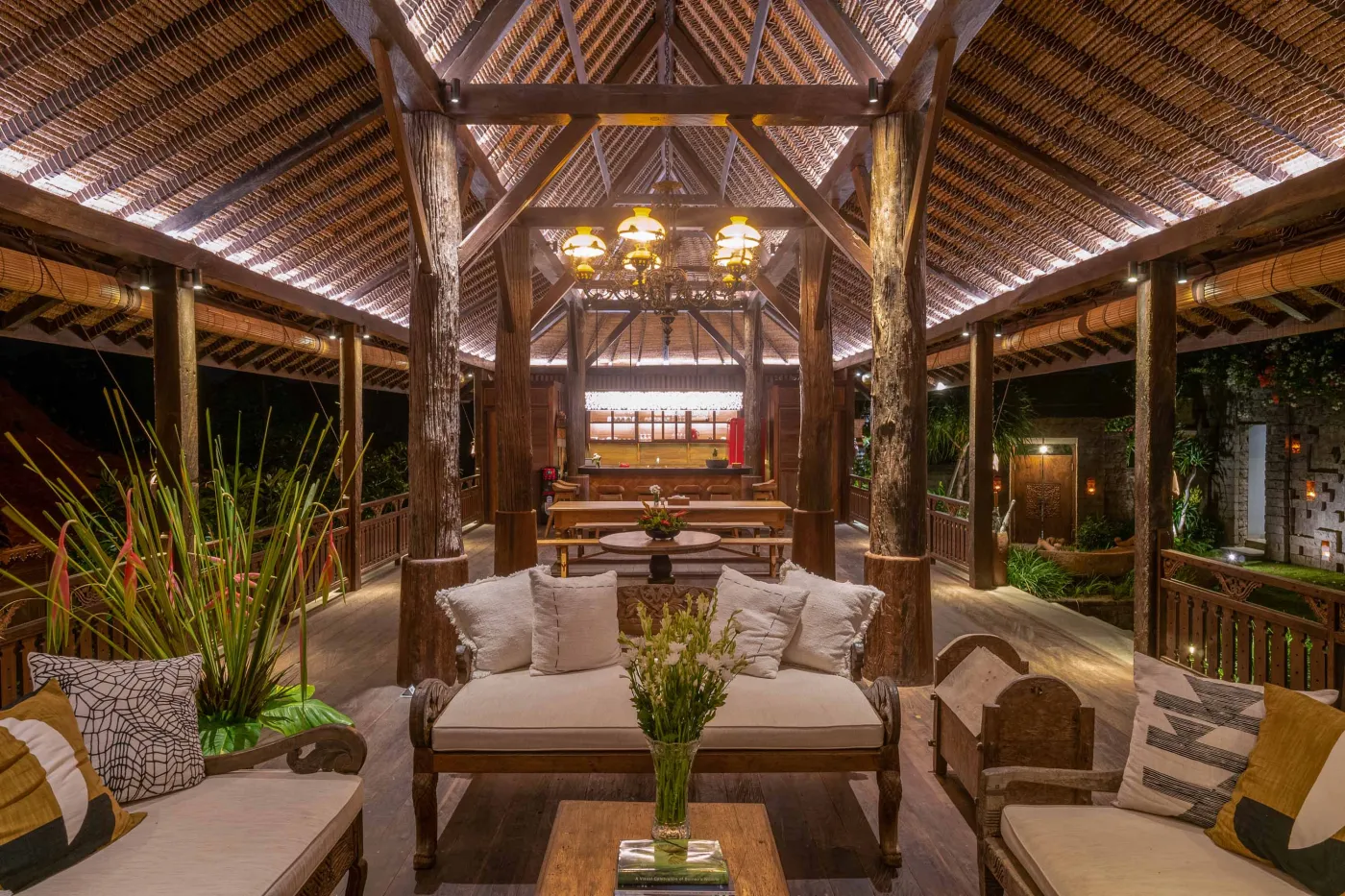Open-air tropical resort bar with carved wooden counter, thatched roof, backlit liquor shelves, fresh fruits, and surrounding lush greenery. - Bali Villas