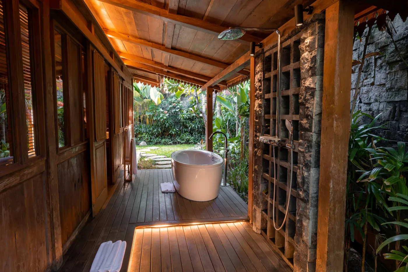 Open-air tropical resort bar with carved wooden counter, thatched roof, backlit liquor shelves, fresh fruits, and surrounding lush greenery. - Bali Villas