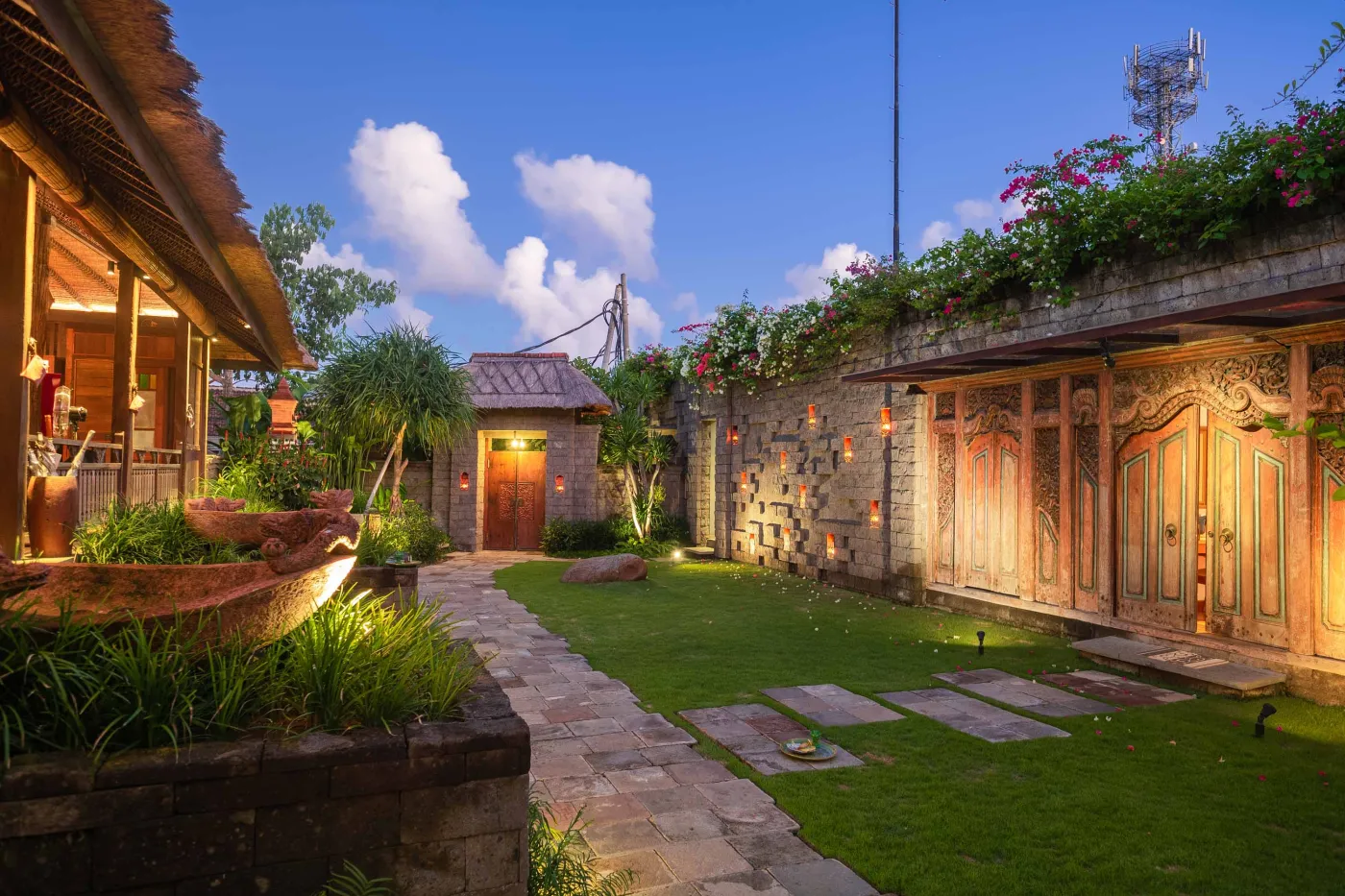 Open-air tropical resort bar with carved wooden counter, thatched roof, backlit liquor shelves, fresh fruits, and surrounding lush greenery. - Bali Villas