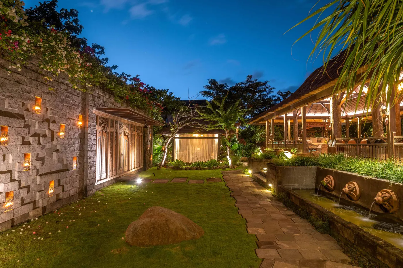 Open-air tropical resort bar with carved wooden counter, thatched roof, backlit liquor shelves, fresh fruits, and surrounding lush greenery. - Bali Villas