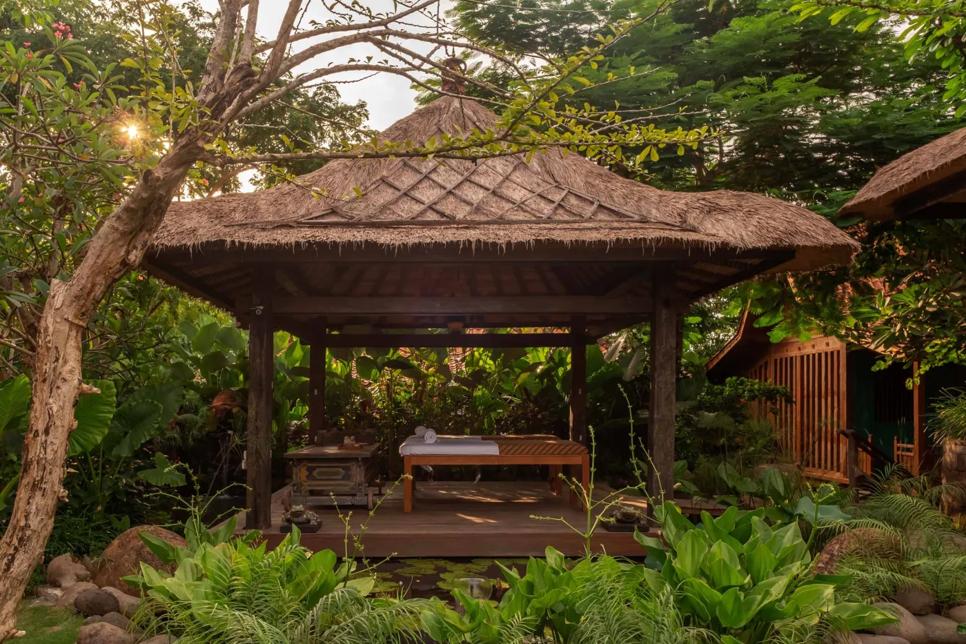 Open-air tropical resort bar with carved wooden counter, thatched roof, backlit liquor shelves, fresh fruits, and surrounding lush greenery. - Bali Villas