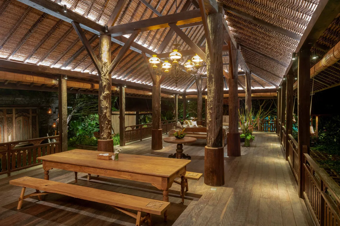 Open-air tropical resort bar with carved wooden counter, thatched roof, backlit liquor shelves, fresh fruits, and surrounding lush greenery. - Bali Villas