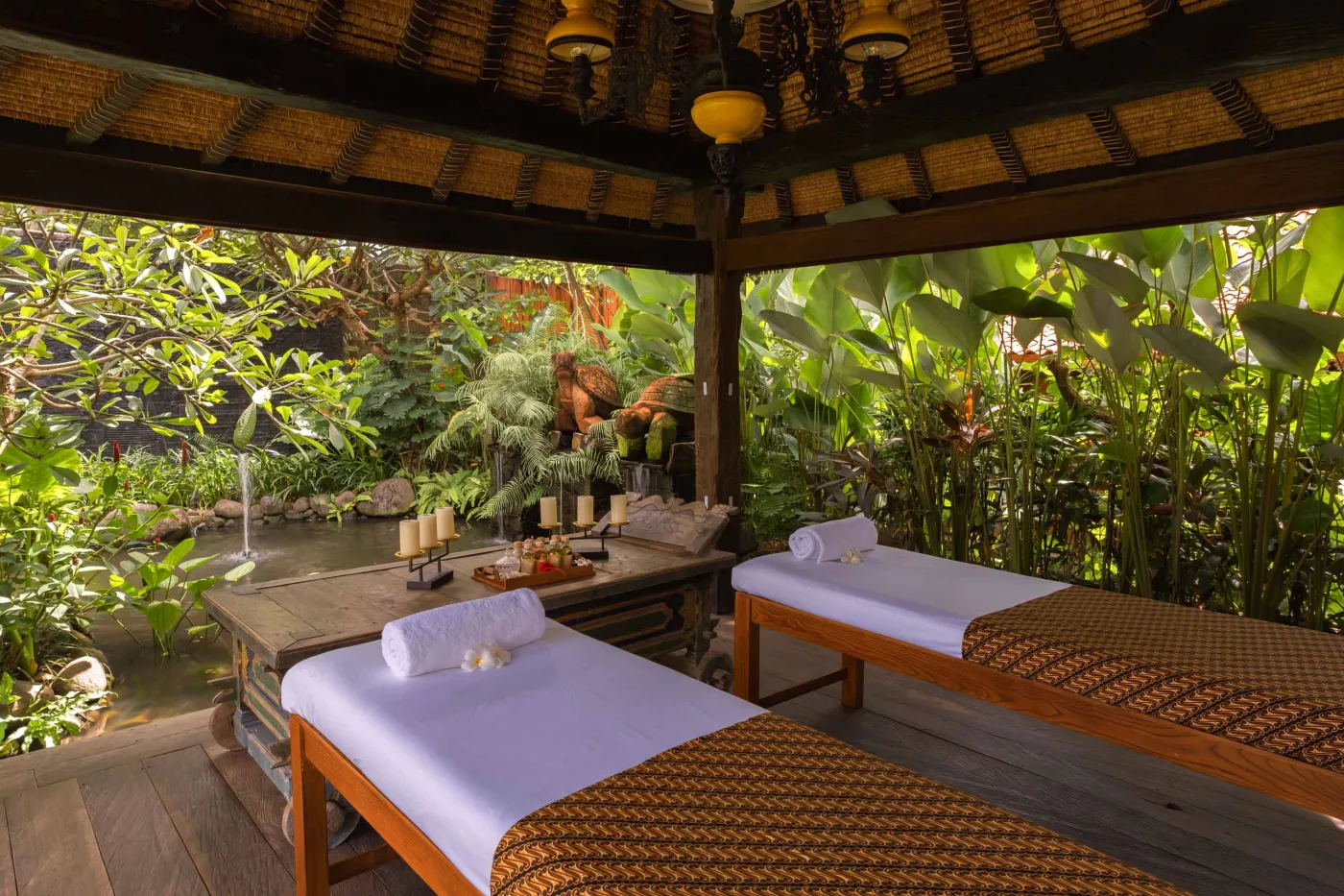 Open-air tropical resort bar with carved wooden counter, thatched roof, backlit liquor shelves, fresh fruits, and surrounding lush greenery. - Bali Villas