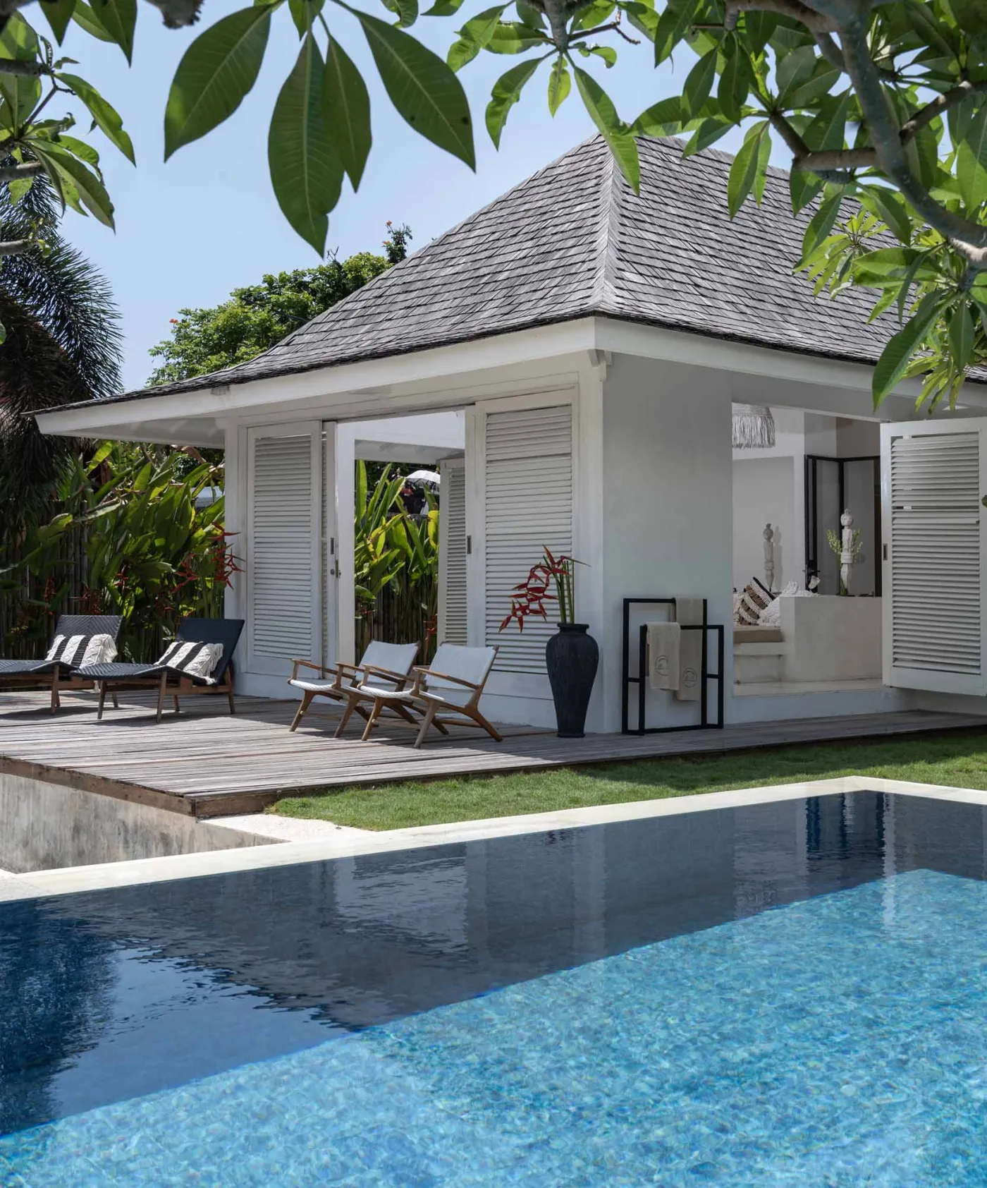 Spacious open-air Balinese villa lounge with white columns, tropical greenery, hanging hammocks, and carved wooden furniture. - Bali Villas