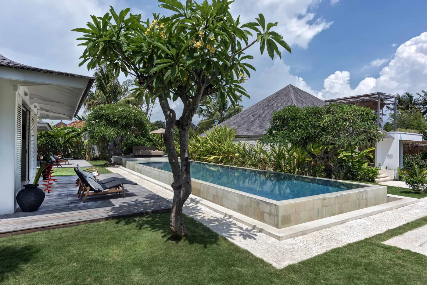 Spacious open-air Balinese villa lounge with white columns, tropical greenery, hanging hammocks, and carved wooden furniture. - Bali Villas