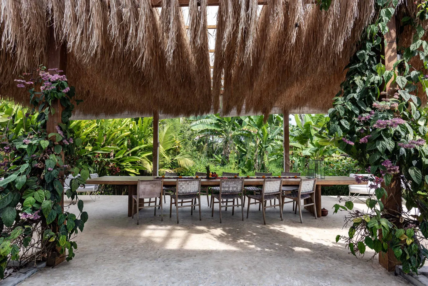 Spacious open-air Balinese villa lounge with white columns, tropical greenery, hanging hammocks, and carved wooden furniture. - Bali Villas