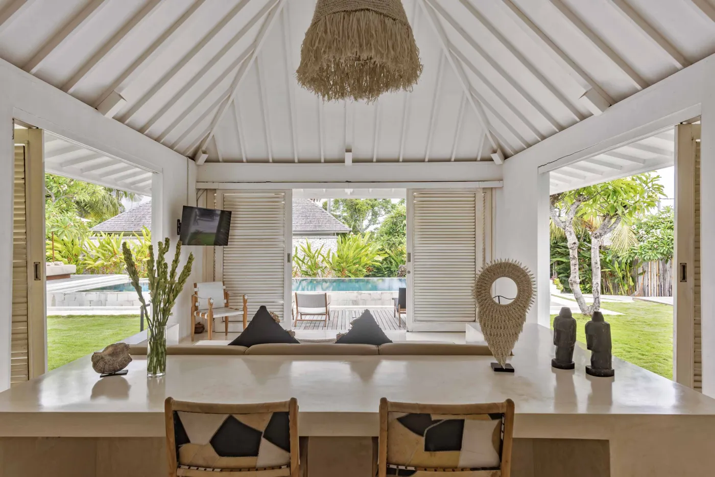 Spacious open-air Balinese villa lounge with white columns, tropical greenery, hanging hammocks, and carved wooden furniture. - Bali Villas