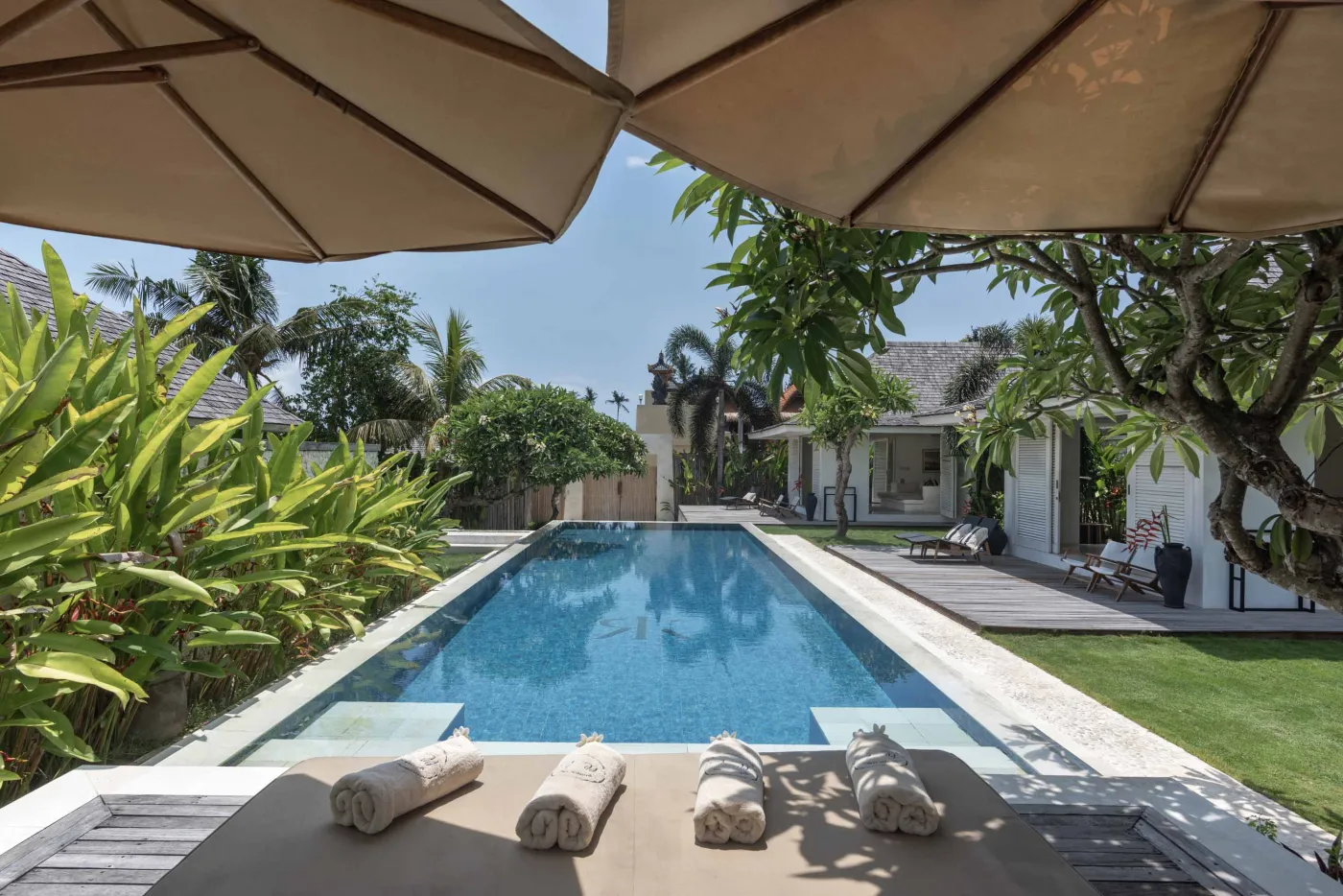 Spacious open-air Balinese villa lounge with white columns, tropical greenery, hanging hammocks, and carved wooden furniture. - Bali Villas