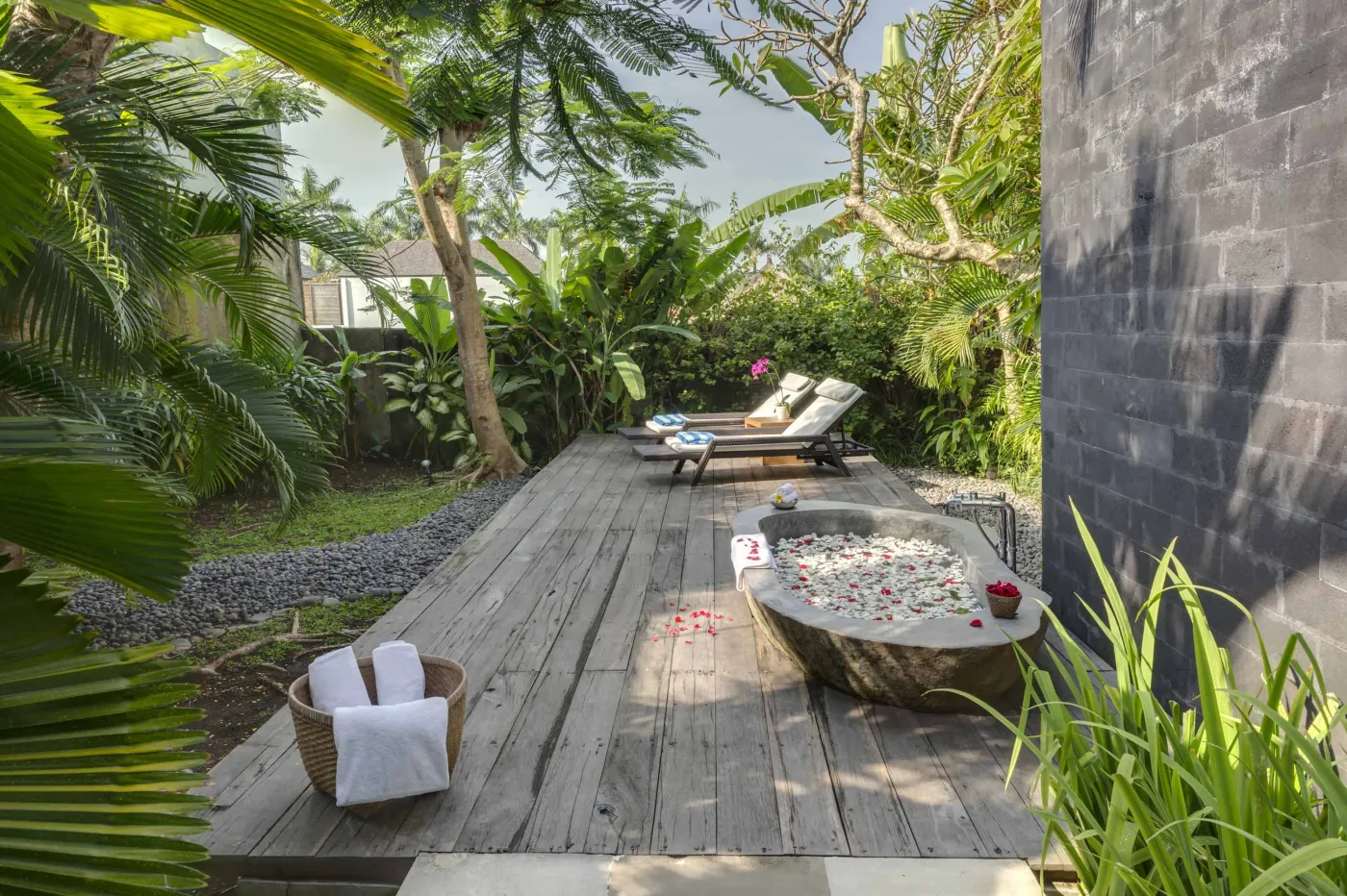 Serene tropical outdoor deck in a Bali villa featuring a stone bathtub filled with rose petals, lounge chairs, towels, and lush greenery. - Bali Villas