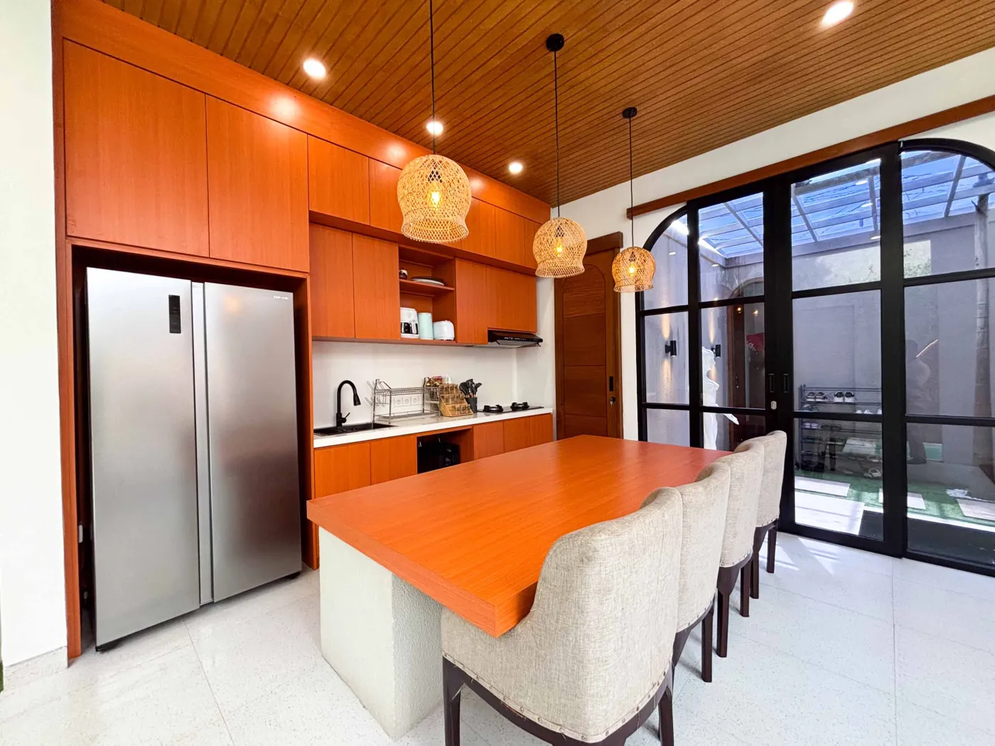 Modern tropical villa kitchen and dining area with orange cabinets, wooden ceiling, pendant lights, and outdoor pool view featuring a statue. - Bali Villas