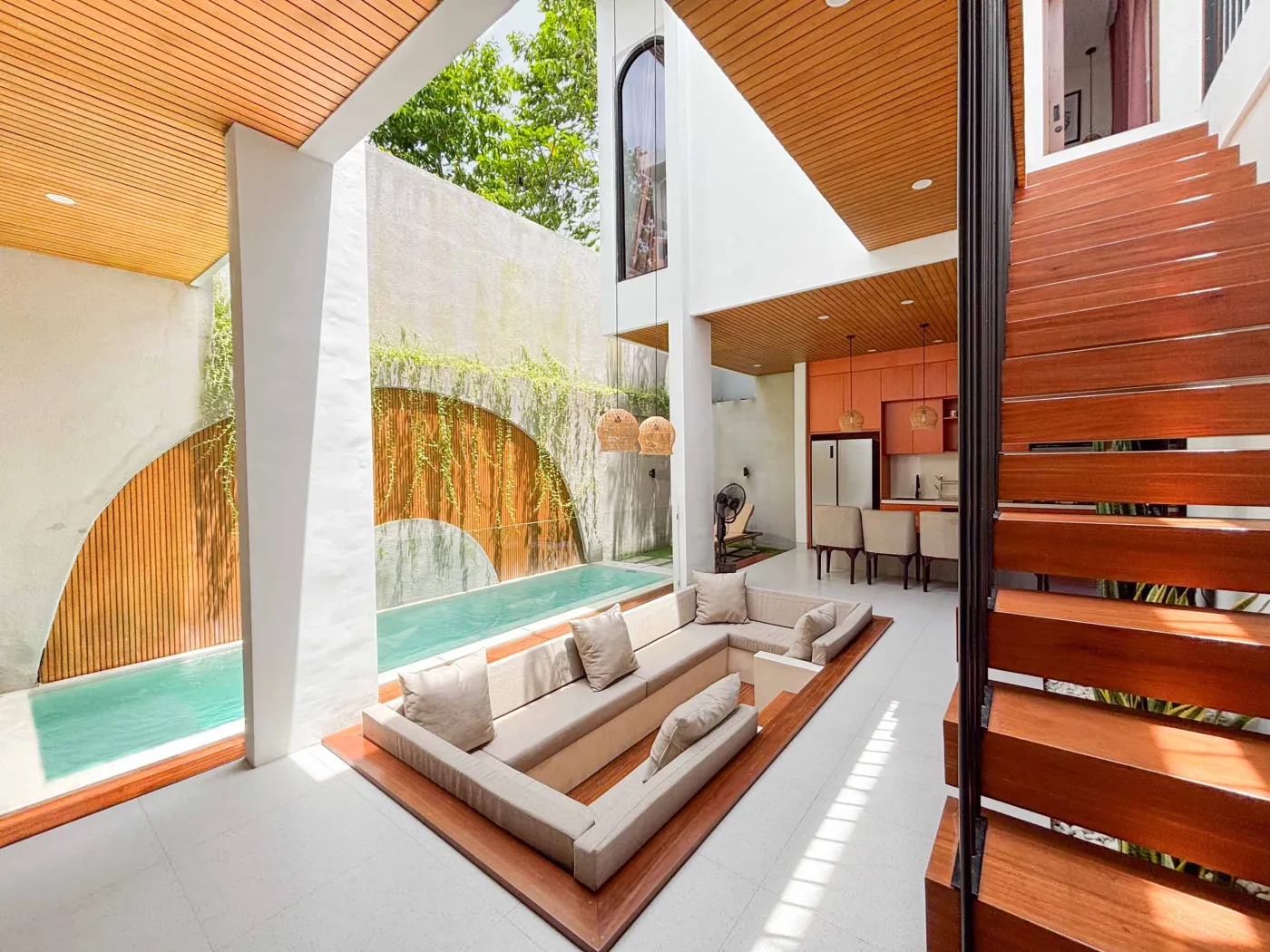 Modern tropical villa kitchen and dining area with orange cabinets, wooden ceiling, pendant lights, and outdoor pool view featuring a statue. - Bali Villas