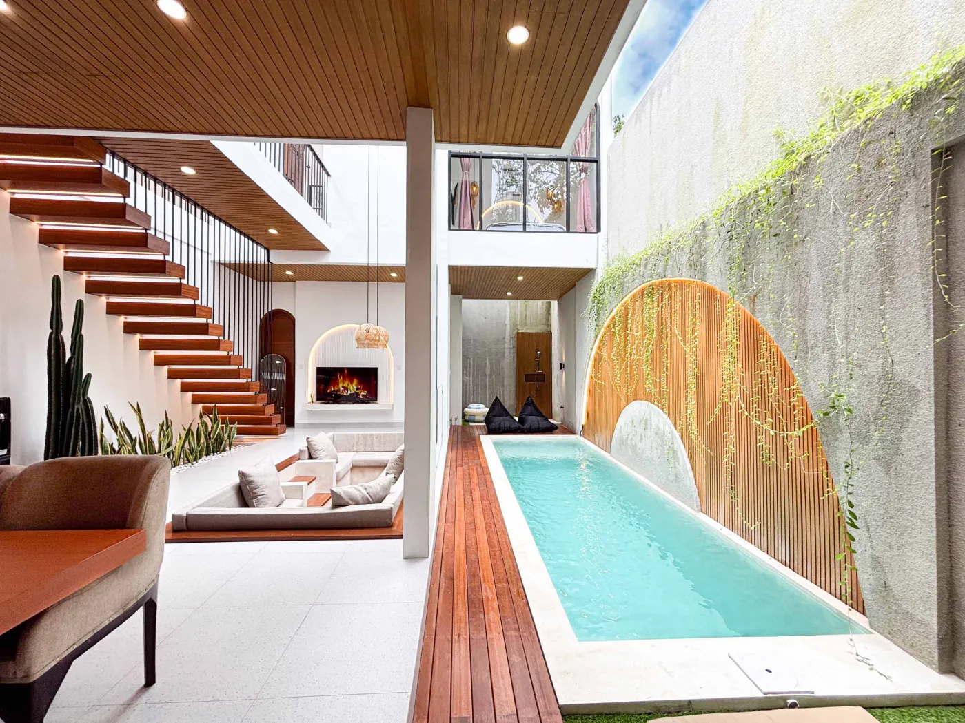 Modern tropical villa kitchen and dining area with orange cabinets, wooden ceiling, pendant lights, and outdoor pool view featuring a statue. - Bali Villas