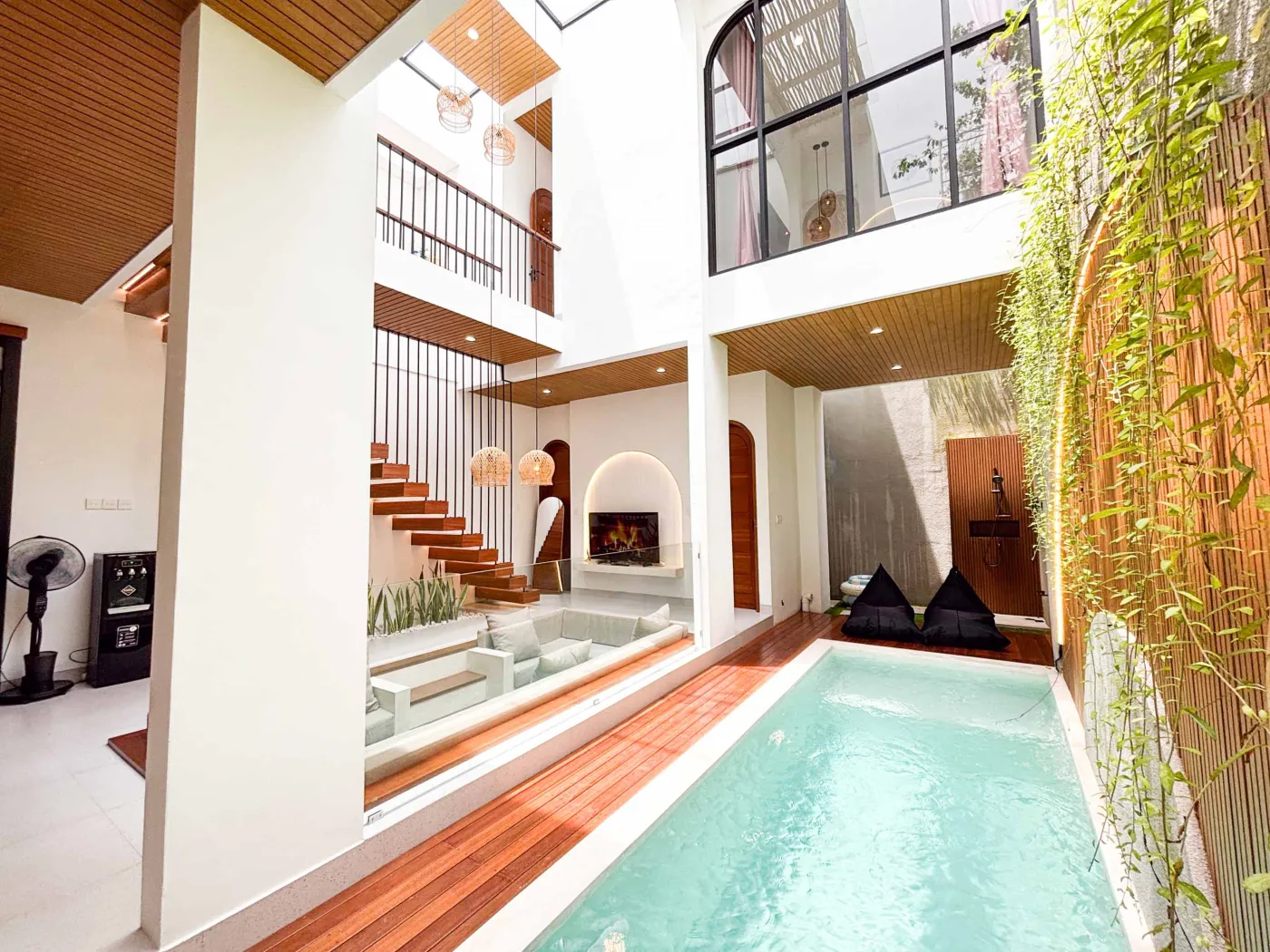 Modern tropical indoor atrium with a serene lap pool, wooden decking, cushioned lounge seating, and lush greenery views through arched windows and bamboo screens. - Bali Villas