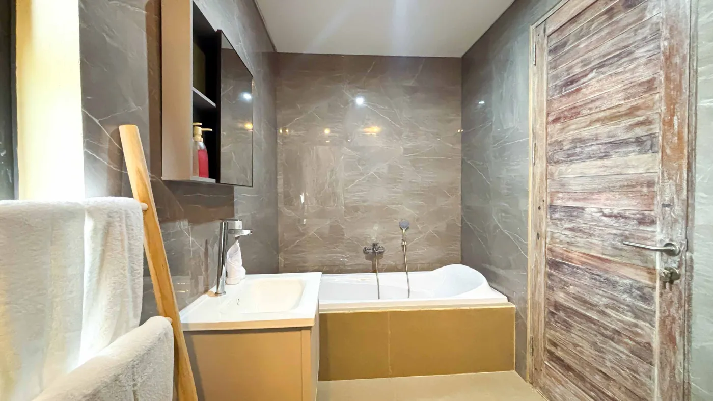 Modern bathroom with gray tile walls, corner soaking tub, wooden vanity sink, towel ladder, and rustic reclaimed wood door. - Bali Villas