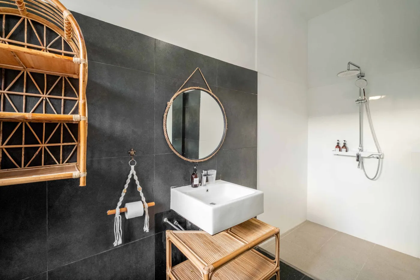 Modern tropical bathroom with dark tiled walls, rattan shelf and bamboo accents, oval rope mirror, white sink, bidet toilet, and window view of poolside greenery. - Bali Villas
