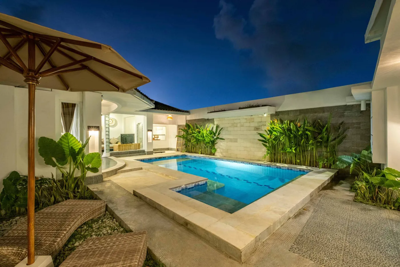 Modern tropical bathroom with dark tiled walls, rattan shelf and bamboo accents, oval rope mirror, white sink, bidet toilet, and window view of poolside greenery. - Bali Villas