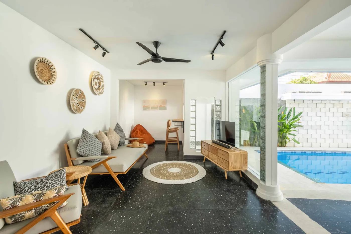 Modern tropical bathroom with dark tiled walls, rattan shelf and bamboo accents, oval rope mirror, white sink, bidet toilet, and window view of poolside greenery. - Bali Villas